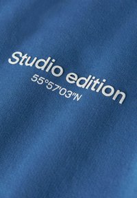 Niebieska tkanina z białym napisem "Edytcja studia 55°57'03"N" nadrukowanym na niej.