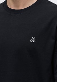 Gros plan d'un t-shirt noir à col rond avec de petites lettres blanches "M O'P" imprimées sur la partie gauche de la poitrine.