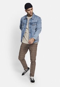 Jeansjacke mit hellblauer Waschung, Kettenakzent an der Tasche, über einem beigen Hemd, kombiniert mit braunen karierten Hosen und schwarzen Sneakers.