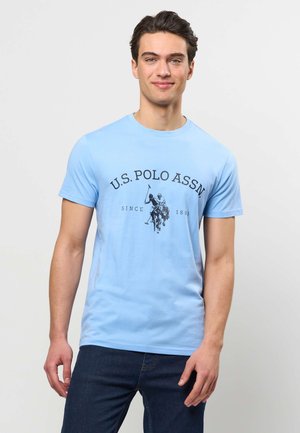 Junger Mann trägt ein hellblaues U.S. Polo Assn. T-Shirt mit dunkelblauen Jeans und steht vor einem schlichten hellen Hintergrund.