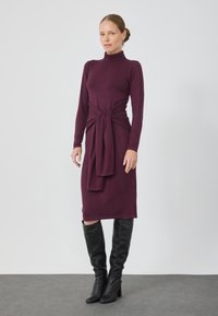 Langemouwen, hoge hals, bordeauxrood jurk met een geknoopt taille-detail. Gekocht met hoge zwarte leren laarzen, met een gladde textuur en een aangesloten silhouet.