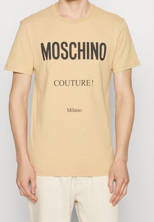 T-shirt beige à manches courtes avec texte noir "MOSCHINO" et texte plus petit "COUTURE !" et "Milano", porté par une personne debout.