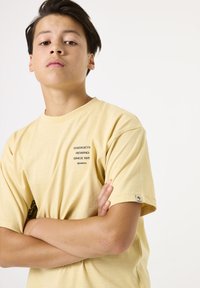 Camiseta de algodón amarilla con mangas cortas, que presenta estampados de logo en el pecho y el costado. Textura suave y corte relajado. Etiqueta de marca pequeña en la manga.