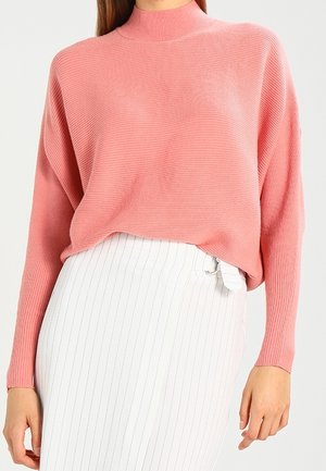 Femme portant un pull rose côtelé à manches longues, rentré dans une jupe blanche taille haute à fines rayures verticales et avec une boucle sur le côté.