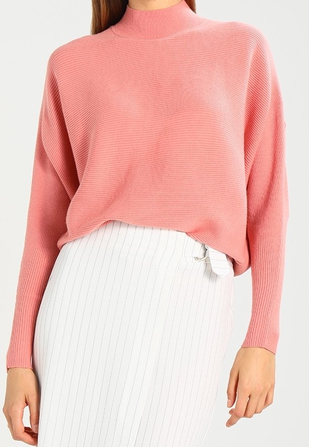 Femme portant un pull rose côtelé à manches longues, rentré dans une jupe blanche taille haute à fines rayures verticales et avec une boucle sur le côté.