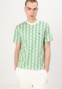 Tricou cu mânecă scurtă în bej deschis, cu un model geometric verde. Material moale, guler normal, cu un detaliu discret al logo-ului pe piept.