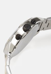 Montre en métal argenté avec un boîtier rond, des poussoirs noirs et un bracelet. Le design présente une finition lisse et une surface polie.