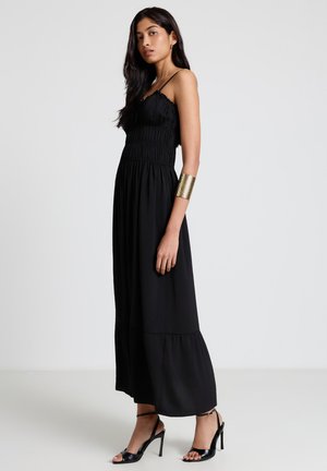 Frau steht in einem schwarzen, ärmellosen Maxikleid, trägt schwarze Sandalen mit hohem Absatz und einen breiten goldenen Manschettenarmreif am linken Arm.