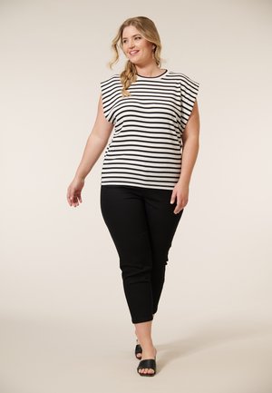 Model loopt zelfverzekerd, gekleed in een gestreepte top en zwarte broek, met een lichte achtergrond die haar ontspannen, stijlvolle uitstraling benadrukt.