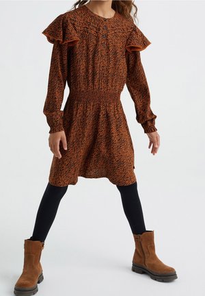 Mädchen trägt ein langärmeliges Kleid mit Rost- und Schwarzmuster, Rüschen an den Schultern, schwarze Leggings und braune Wildleder-Stiefeletten.