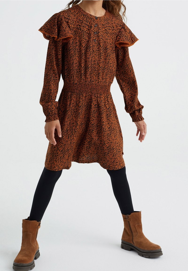 Niña con un vestido de manga larga color óxido y negro con estampado y hombros con volantes, leggins negros y botines de ante marrones.