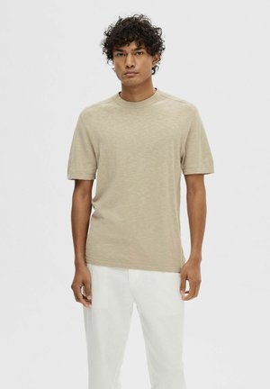 T-shirt beige a maniche corte realizzata in materiale morbido, con collo rotondo e vestibilità rilassata, abbinata a pantaloni bianchi. Design minimalista.