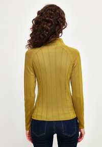 Senfgelbes Langarmoberteil mit vertikal gerippter Textur, hohem Kragen und figurbetontem Design. Kombiniert mit dunkelblauen Jeans.