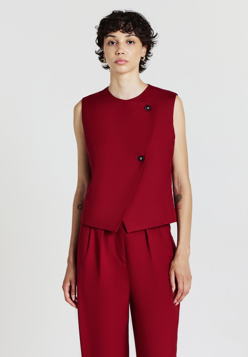 Donna con i capelli ricci e corti, che indossa un top rosso senza maniche con due bottoni neri e pantaloni rossi plissettati, su sfondo bianco.