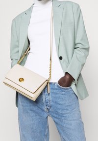 Sac en bandoulière texturé couleur crème avec une chaîne dorée et un fermoir, complété par un blazer vert clair et un turtleneck blanc.