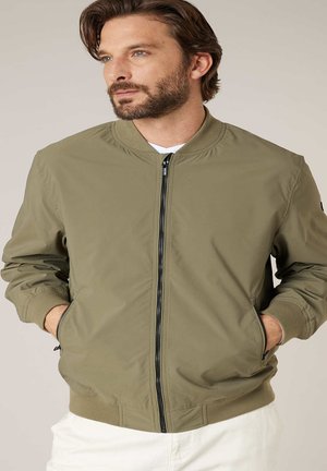 Man met baard, gekleed in een olijfgroene bomberjack met ritssluiting en handen in de zakken, kijkt lichtjes opzij tegen een effen achtergrond.