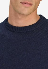 Marineblauwe gebreide trui met een geribde ronde halslijn, die een zachte textuur en een naadloos ontwerp laat zien, met de nadruk op warmte en comfort.