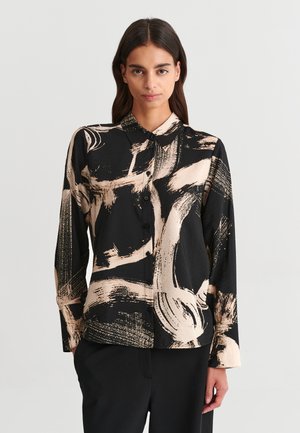 Femme aux longs cheveux foncés portant une chemise noire boutonnée avec des motifs abstraits beige de coups de pinceau et un pantalon noir, debout devant un fond uni.