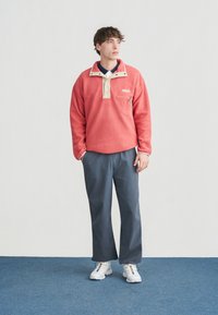 Νέος άνδρας που στέκεται με τα χέρια στις τσέπες, φοράει ροδακινί φλις πουλόβερ, γκρι παντελόνι, λευκά αθλητικά παπούτσια, σε απλό λευκό και μπλε φόντο.