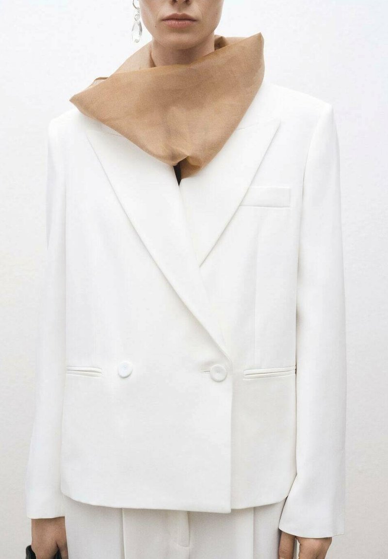 Bílý dvouřadý blazer s volným střihem, textured látkou a dvěma knoflíky. Nosený s béžovým šátkem kolem krku.