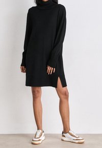 Žena v černých dlouhých rukávech rolákových šatech s bočním rozparkem, v kombinaci s bílými a hnědými teniskami, stojí proti bílé zdi.