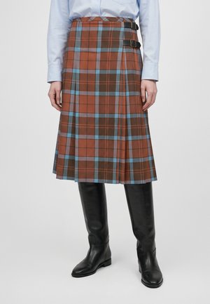 Personne portant un kilt plissé à carreaux rouille et bleus, avec deux sangles à boucle, une chemise blanche et des bottes noires montant jusqu’aux genoux.