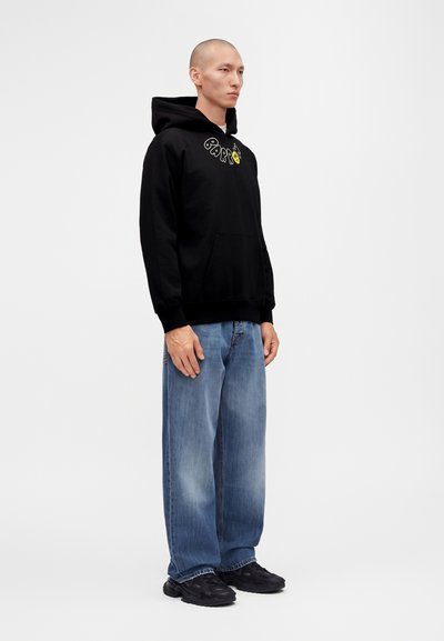 Homme debout portant un sweat à capuche noir avec texte, jean bleu ample et baskets noires sur un fond blanc uni.