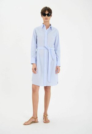 Robe rayée bleu clair avec un devant à boutons, des manches longues et une ceinture nouée à la taille. Portée avec des sandales marron et des lunettes de soleil noires.