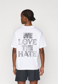 Baltos medvilninės marškinėliai su ryškiu juodu blizgučiu tekstu "MES MYLIME NEAPYKANTĄ" ant nugaros. Apvalus kaklas, trumpomis rankovėmis, laisvas kirpimas.