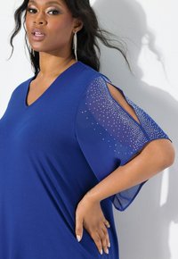 Top blu con scollo a V e maniche corte trasparenti decorate con borchie argentate; tessuto liscio con vestibilità rilassata, che mette in evidenza una delicata drappeggiatura.