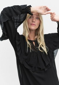 Blondynka w czarnej bluzce z falbanami i prześwitującymi, szerokimi rękawami, z uniesionymi ramionami blisko twarzy na jednolitym jasnym tle.