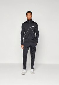 Svart sportoverall i slät tyg; har vita ränder på ärmar och byxben, med en jacka med dragkedja och logotyp på bröstet.