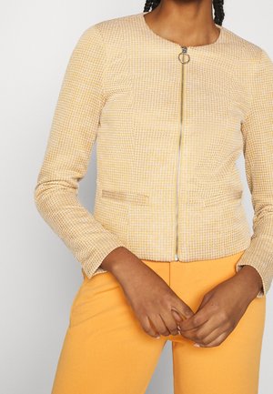 Woman wearing a yellow textured zip-up jacket with ring pull and bright yellow pants, hands clasped at waist.