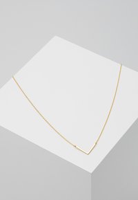 Goldene Halskette mit einer dünnen Kette und einem V-förmigen Anhänger, der eine glatte Textur und ein minimalistischem Design auf einem hellgrauen Hintergrund aufweist.