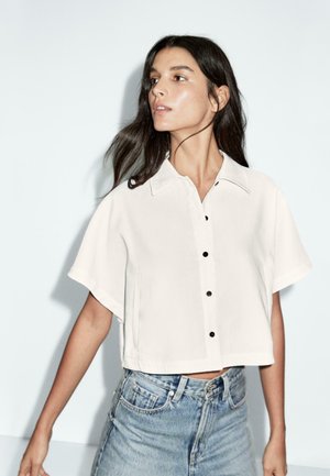 Femme aux longs cheveux noirs portant une chemise blanche à manches courtes boutonnée et un jean bleu taille haute, regardant vers la gauche sur un fond uni.