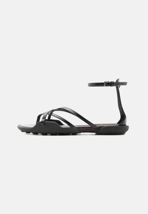 Sandale plate noire avec de fines brides croisées sur les orteils et une bride de cheville à boucle, avec une semelle légèrement texturée pour une meilleure adhérence.