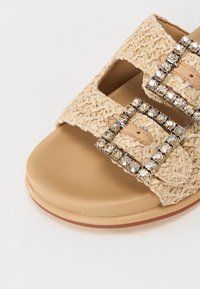 Beige vevd sandal med to brede stropper, dekorert med rhinestones, og en glatt beige fotpute for komfort. Flat såledesign.