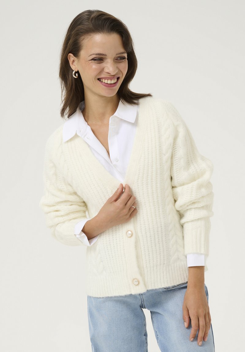Cárdigan de punto crema con escote en V, que presenta un patrón de trenzas y dos cierres de botón, llevado sobre una camisa blanca de cuello y jeans claros.