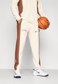 Nike kelnaitės kreminės spalvos su rudos spalvos šoninėmis juostomis, su "The Town" logotipu ir NBA emblema. Dėvimos su baltomis sportbačiais, laikant krepšinio kamuolį.