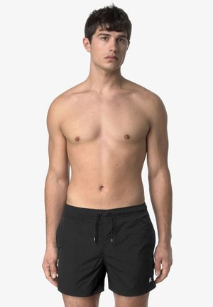 Giovane uomo con capelli corti e scuri, senza maglietta, indossa pantaloncini da bagno neri con coulisse, in piedi davanti a uno sfondo grigio chiaro.