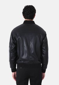 Giubbotto bomber in pelle nera con colletto e polsini a costine, texture liscia e design aderente. Cuciture visibili lungo la schiena e le spalle.
