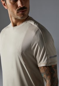 T-shirt de sport beige clair en tissu lisse avec un motif en grille texturé sur les épaules. Design à manches courtes avec des détails d'accent subtils.