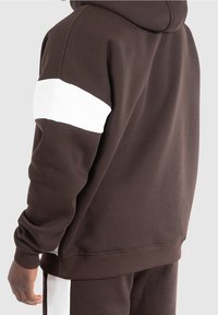 Hoodie marron avec une bande blanche sur le bras, fabriqué en tissu doux. Il dispose d'une poche kangourou et de poignets et un ourlet côtelés.