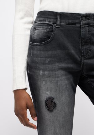 Black denim jeans featuring a faded wash, distressed patches, and intricate black embroidery. Includes standard front pockets and a button closure.