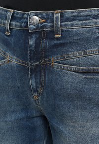 Jean en denim bleu foncé avec coutures visibles, bouton en métal, passants pour ceinture et petite étiquette "CLOSED" près de la fermeture éclair.