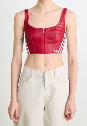 Kobieta ubrana w czerwoną, bezrękawnikową bluzkę typu crop top z zamkiem z przodu i białymi paskami po bokach, zestawioną z jasnobeżowymi spodniami z wysokim stanem.
