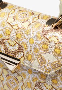 Rakennepintainen käsilaukku, jossa monimutkaiset keltaiset ja ruskeat kukkakuvioinnit, kullanvärisillä metalliosilla ja jämäkällä muotoilulla.