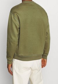 Vue de dos d'une personne portant un sweat-shirt à manches longues vert olive et un pantalon écru sur un fond uni.