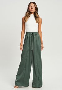 Groene satijnen broek met wijde pijpen en een koord in de taille, gecombineerd met een fitted witte mouwloze top. Gladde textuur en vloeiende uitstraling.