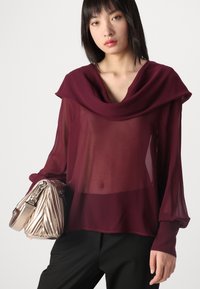Blusa trasparente bordeaux con ampio colletto drappeggiato, maniche lunghe e polsini aderenti. Abbinata a una borsa plissettata argento e pantaloni neri.